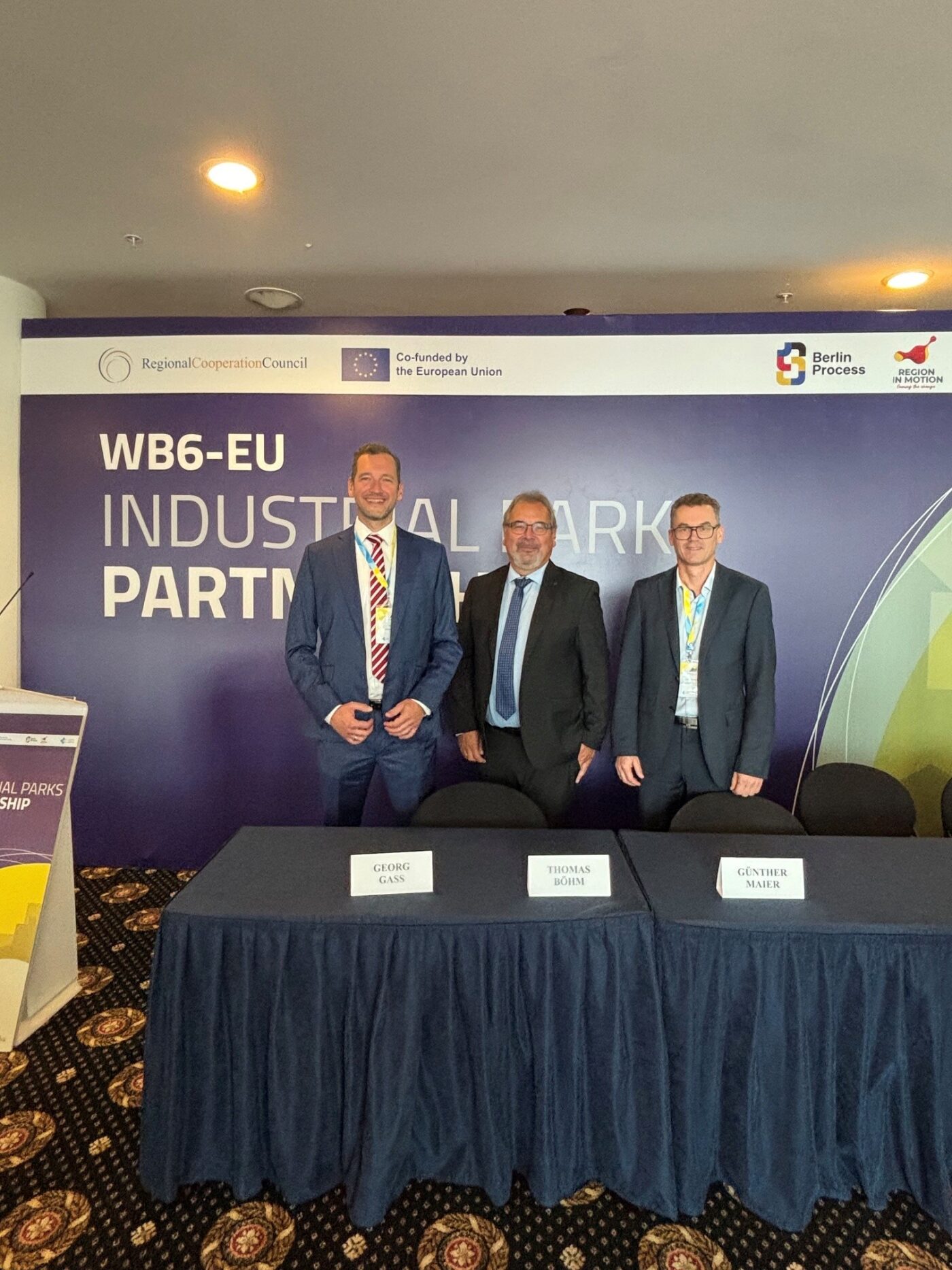 Drei Männer stehen hinter einem Tisch bei der WB6-EU Industrialpark-Partnerschaft Konferenz mit EU-Logos.