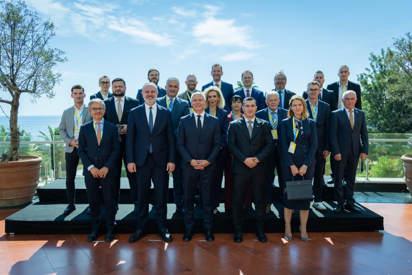 Gruppenfoto von etwa 25 Personen in Business-Anzügen mit Auszeichnungen an einer Küstenanlage mit Meerblick.