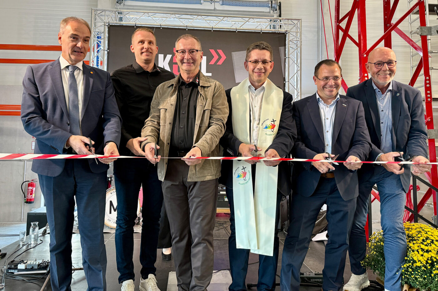 Sechs Männer durchschneiden feierlich ein rotes Band vor einer großen Displaywand in einer Industriehalle zur Eröffnung.