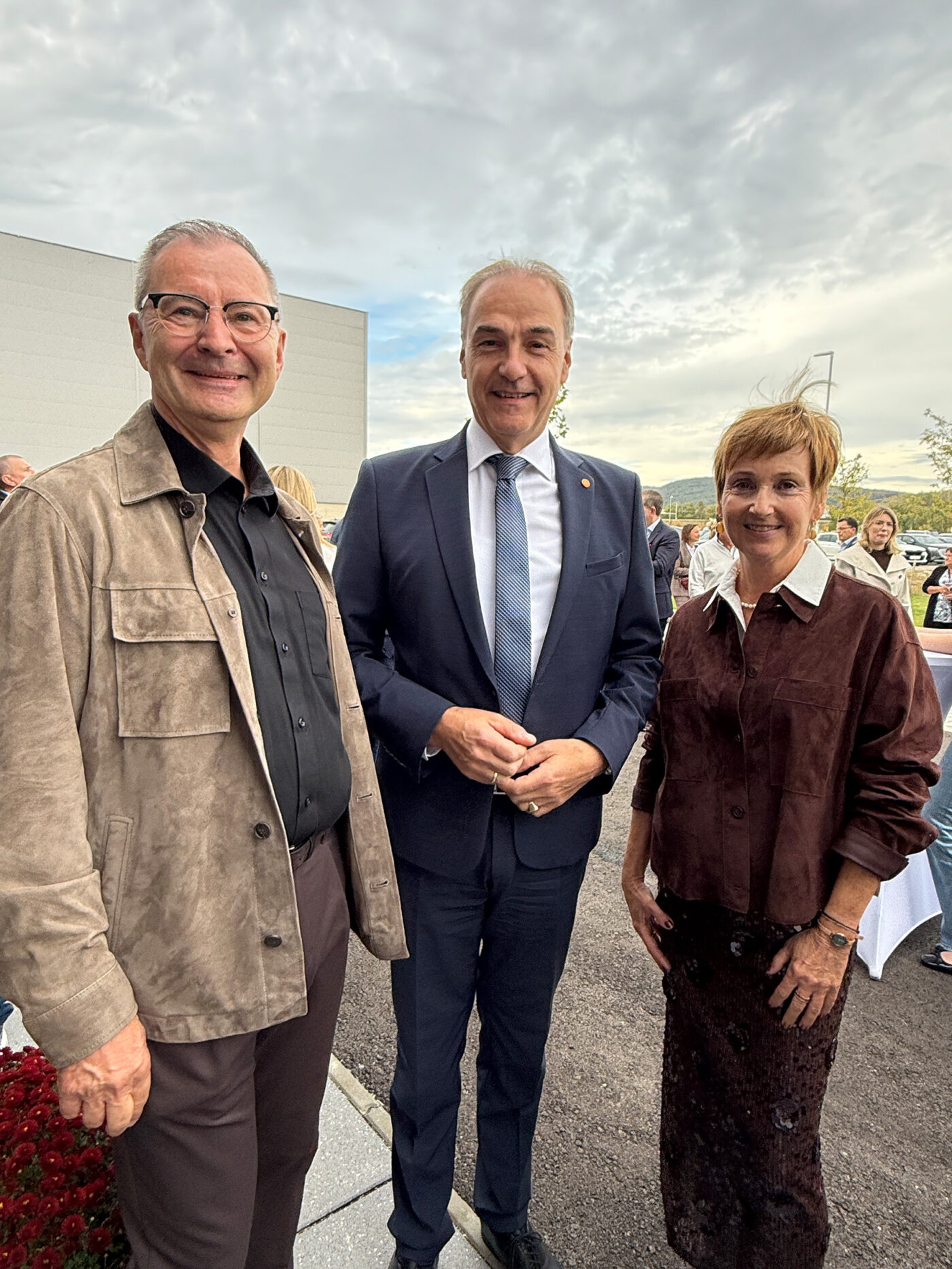 Drei Personen posieren zusammen bei einer Outdoor-Veranstaltung vor einem bewölkten Himmel und Gebäude.