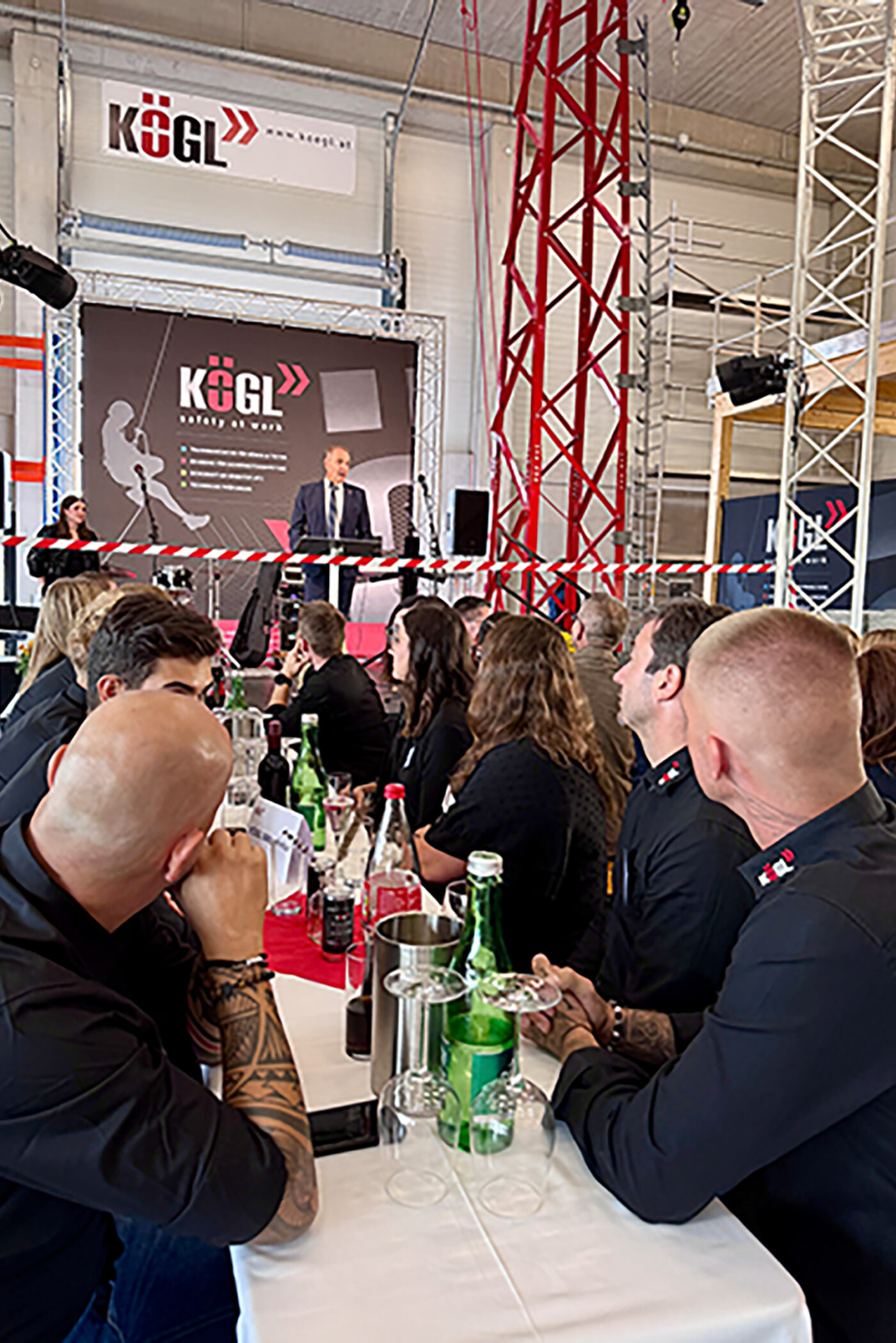 KÖGL Unternehmensveranstaltung mit Redner auf Bühne, Publikum am Tisch mit Getränken, moderne Industriehalle mit Trussing.