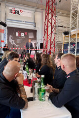 KÖGL Unternehmensveranstaltung mit Redner auf Bühne, Publikum am Tisch mit Getränken, moderne Industriehalle mit Trussing.