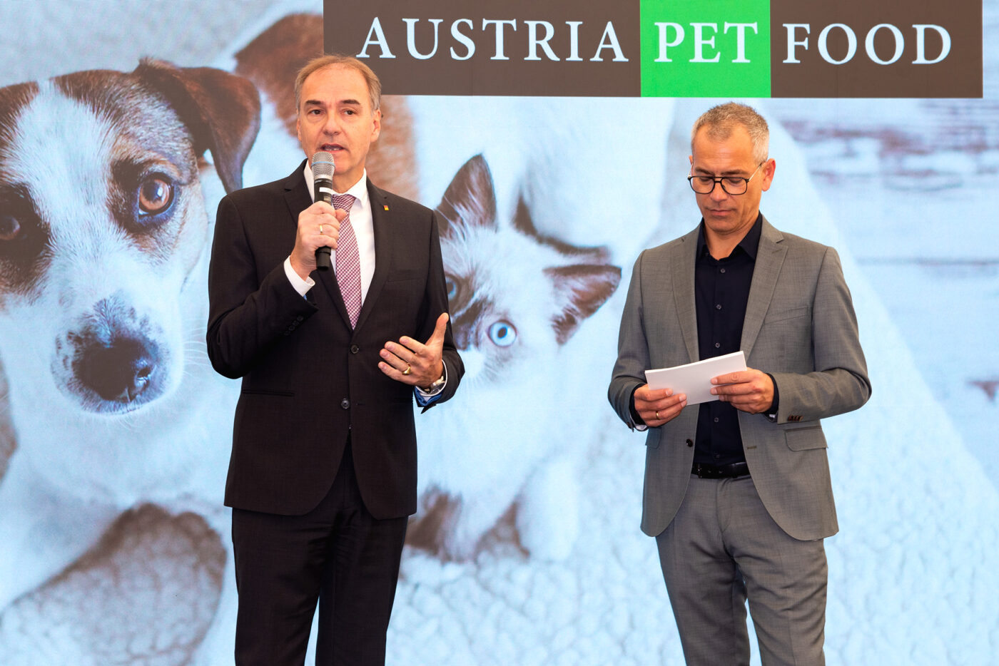 Zwei Geschäftsmänner präsentieren Austria Pet Food auf einer Bühne mit Hundebildern im Hintergrund.