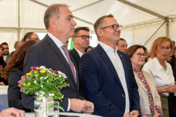 Offizielle Veranstaltung unter Zelt mit mehreren Personen in formaler Kleidung neben Blumengesteck.