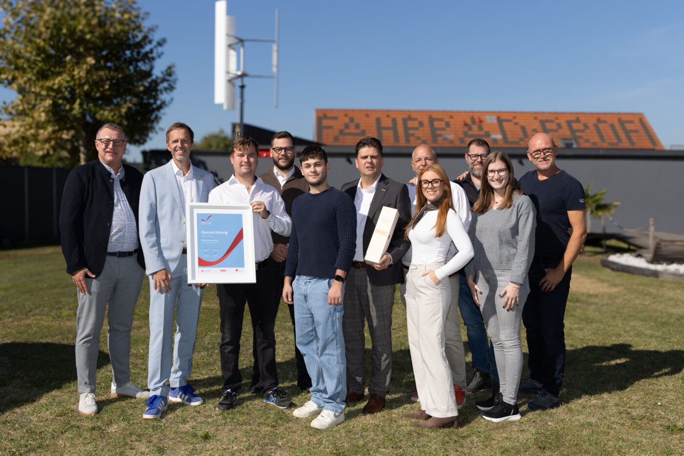 Teamfoto einer Gruppe von zehn Personen auf einer Wiese, die eine Auszeichnung präsentiert, im Hintergrund ein Gebäude mit Solarpanelen. (_r6a8741_innovationspreis-wab)