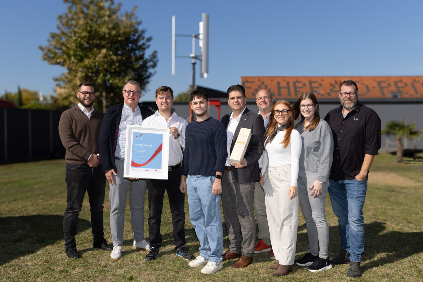 Gruppe von neun Personen präsentiert Auszeichnung vor Gebäude mit Windkraftanlage im Hintergrund. (_r6a8750_innovationspreis-wab)