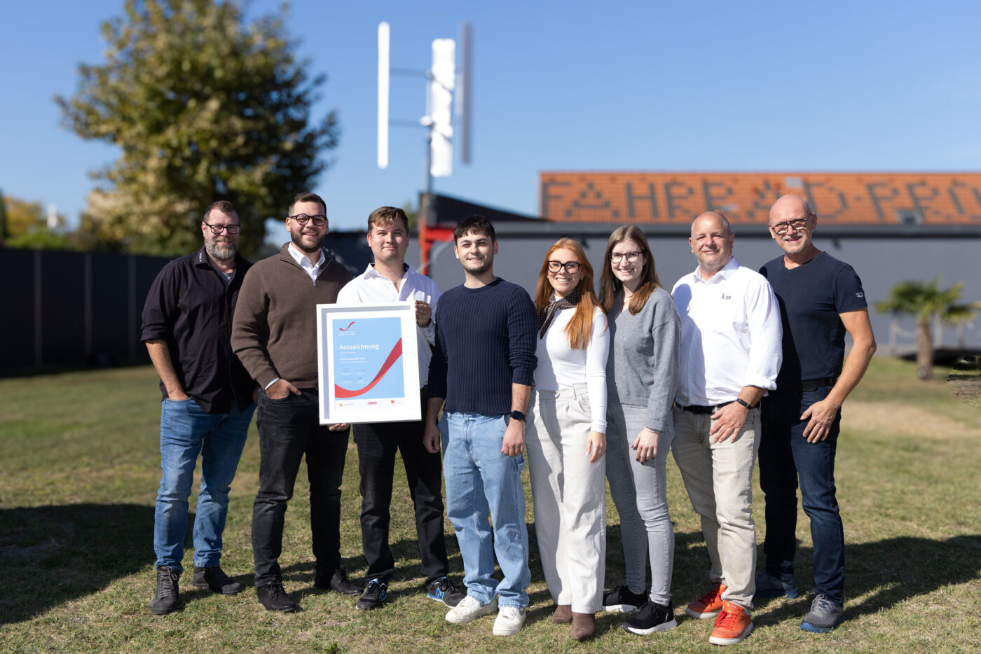 Team mit Zertifikat vor Gebäude, Auszeichnung für Unternehmensleistung, acht Personen posieren stolz mit Urkunde. (_r6a8760_innovationspreis-wab)