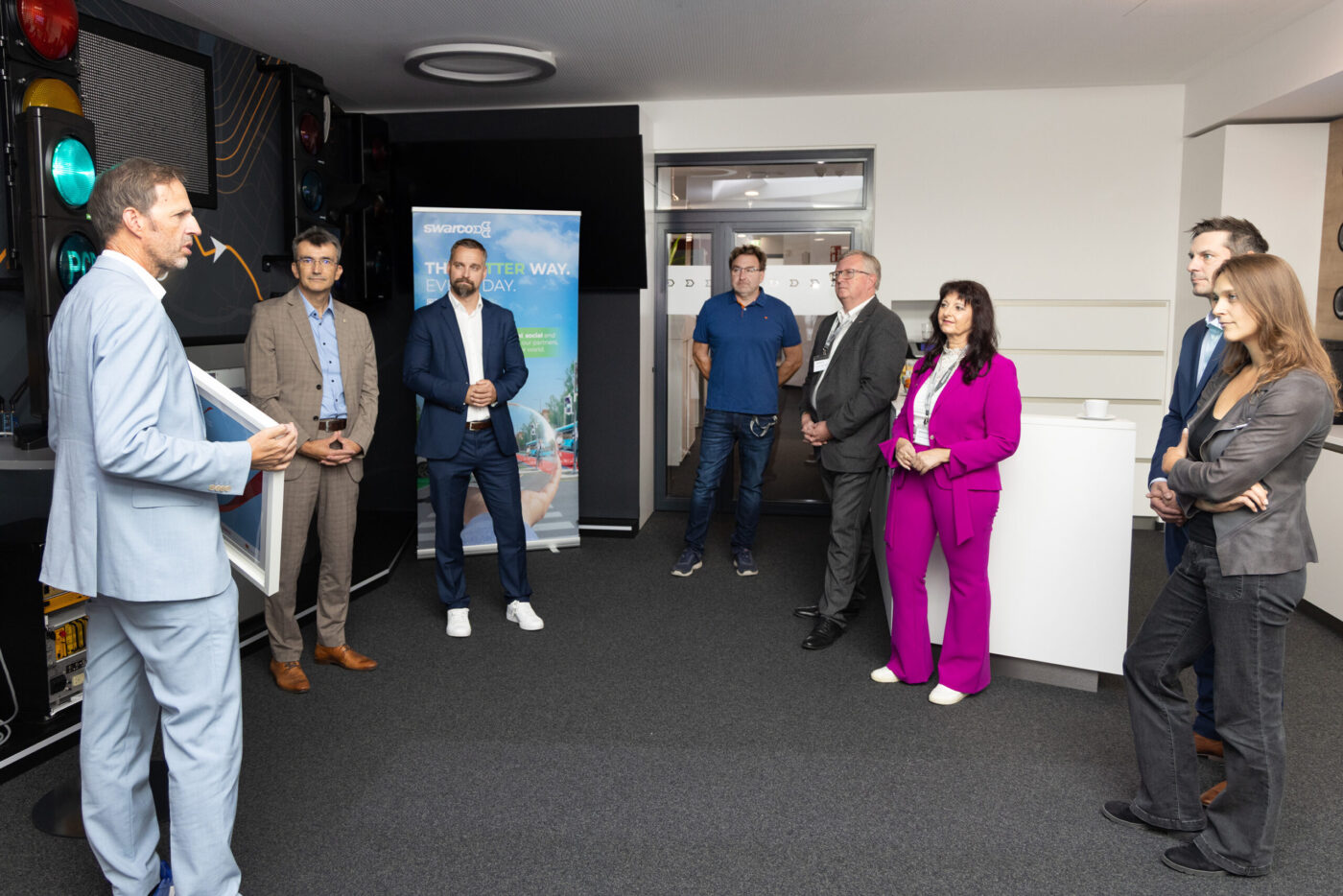 Geschäftspräsentation in modernem Büro: Mann in hellblauem Anzug spricht zu Gruppe von Fachleuten, Swarco Banner sichtbar. (_r6a8798_innovationspreis-wab)