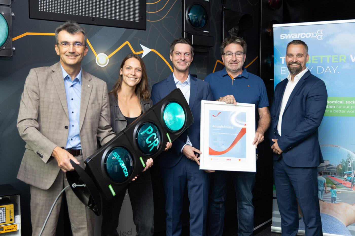 Fünf Personen präsentieren stolz eine Auszeichnung und eine grüne Ampel mit Swarco-Logo vor dunklem Hintergrund. (_r6a8813_innovationspreis-wab)