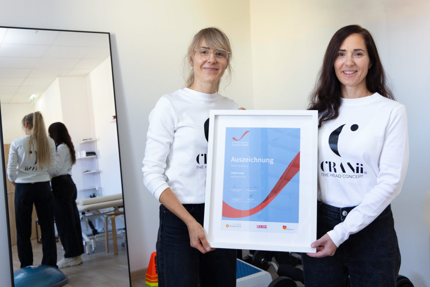 Zwei Frauen in weißen CRANii-Shirts halten eine gerahmte Auszeichnung in einem modernen Büro. (_r6a8848_innovationspreis-wab)