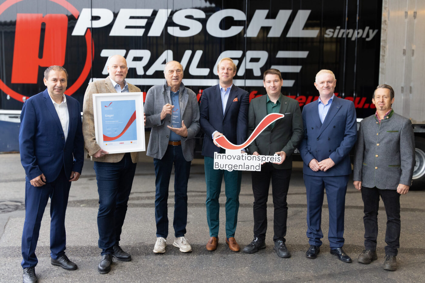 Peischl Trailer gewinnt Innovationspreis Burgenland, sieben Personen halten Auszeichnung und Logo vor Firmengebäude.